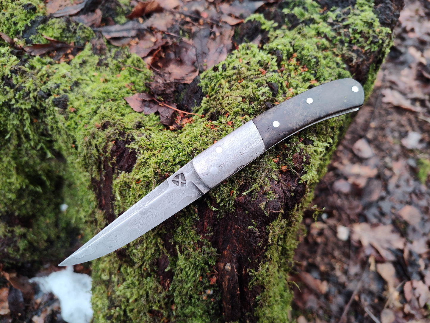 Hand-forged slipjoint folding knife with a pattern-welded blade. Wrought Iron Bolsters with Black Ash Burl Scales. Non-locking UK Legal Carry