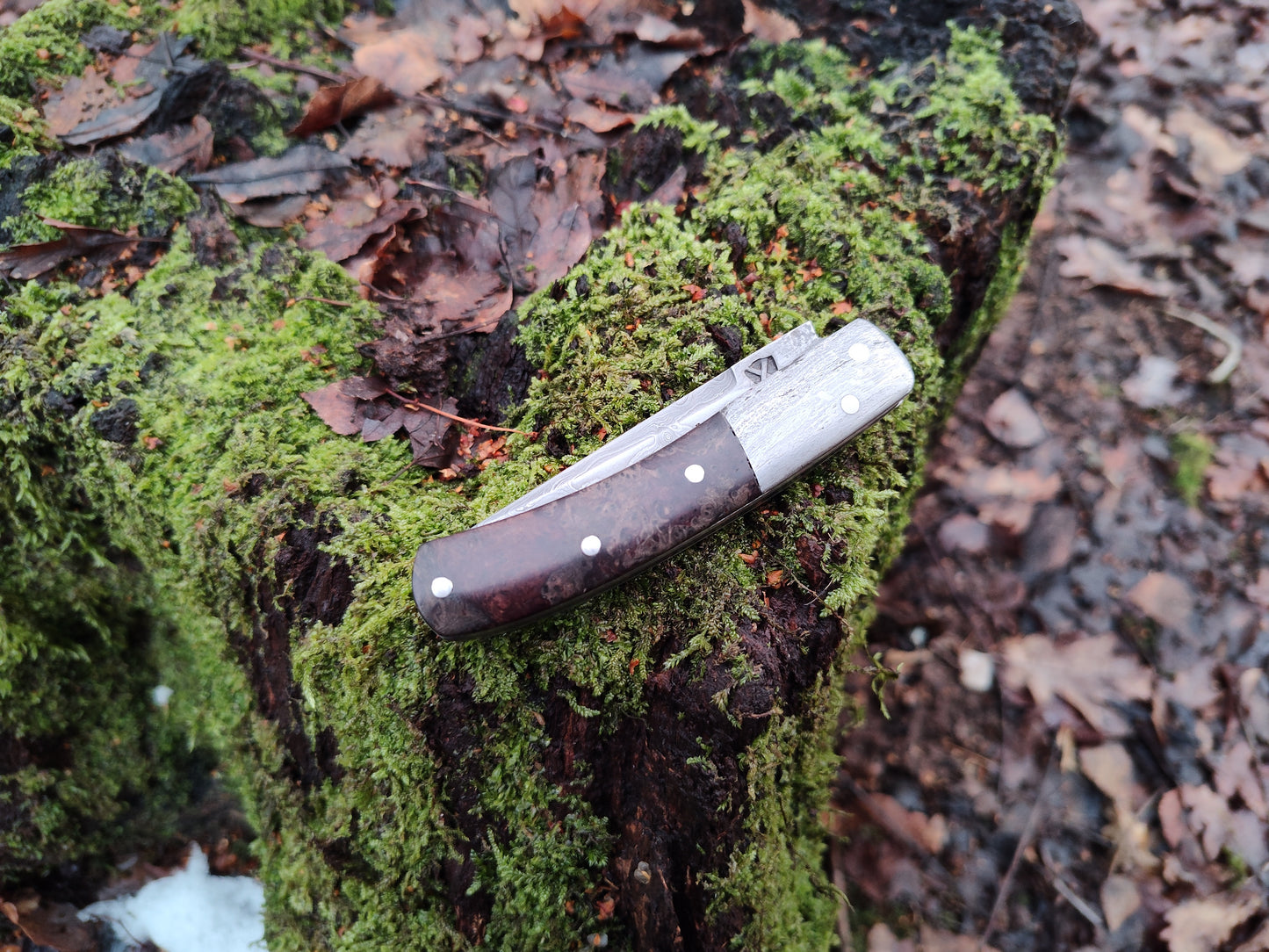 Hand-forged slipjoint folding knife with a pattern-welded blade. Wrought Iron Bolsters with Black Ash Burl Scales. Non-locking UK Legal Carry