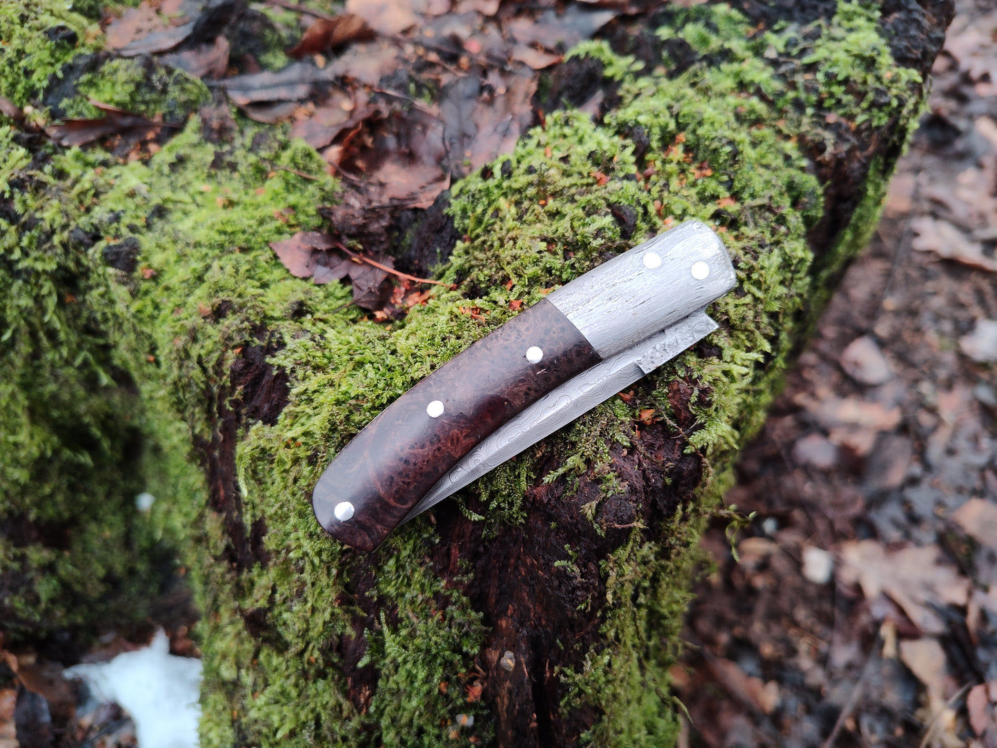 Hand-forged slipjoint folding knife with a pattern-welded blade. Wrought Iron Bolsters with Black Ash Burl Scales. Non-locking UK Legal Carry