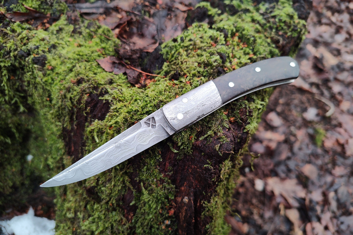 Hand-forged slipjoint folding knife with a pattern-welded blade. Wrought Iron Bolsters with Black Ash Burl Scales. Non-locking UK Legal Carry